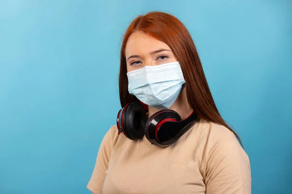年轻的红头发女孩 戴着大大的耳机 戴着蓝色背景的医疗面罩 高质量的照片 — 图库照片