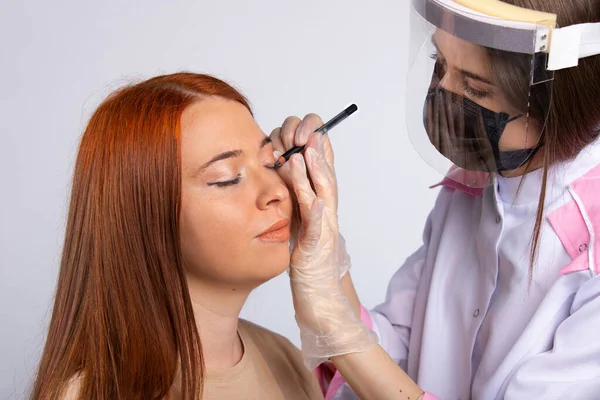 A makeup artist in a robe, mask and gloves makes makeup for a red-haired girl. High quality photo