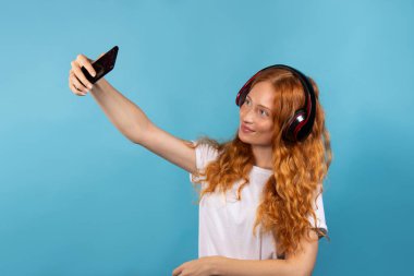 Beyaz tişörtlü güzel kızıl saçlı bir kız kulaklıkla müzik dinliyor ve telefonda selfie çekiyor. Mavi arka planda bir fotoğraf. Yüksek kalite fotoğraf