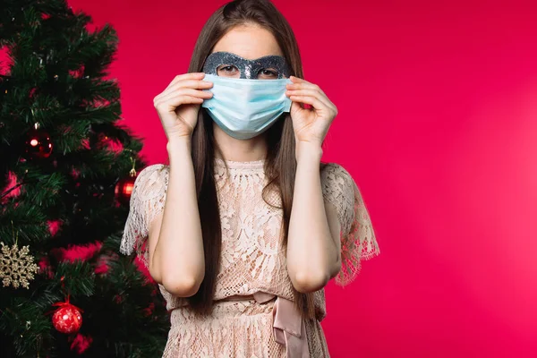 一个戴着狂欢节和医疗面具的女孩 在一棵圣诞树旁边的红色背景上 旁边有一个空旷的侧隙 高质量的照片 — 图库照片