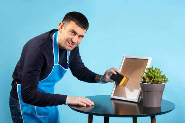 A man in an apron and gloves wipes the mirror and looks at the camera. Photo on a blue background. Gender stereotypes concept. High quality photo