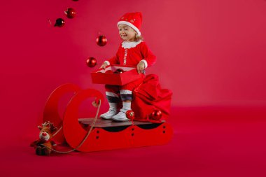 Kırmızı Noel Baba kostümü içinde bir çanta dolusu hediye ve bir kutu Noel süslemesiyle mutlu bir kız. Yan boşluk. Yüksek kalite fotoğraf