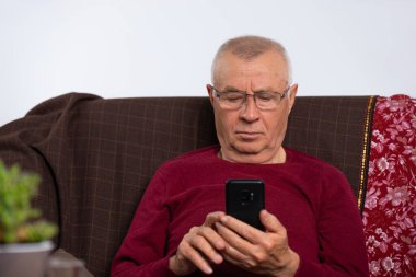 Evde gözlüklü yaşlı adam kanepede oturuyor ve akıllı telefon kullanıyor. Ev ve zaman geçirmek. Yüksek kalite fotoğraf