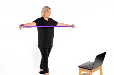 Senior woman rolls a fitness rubber band in her hands while standing in front of a laptop. White background. Online workout concept. High quality photo