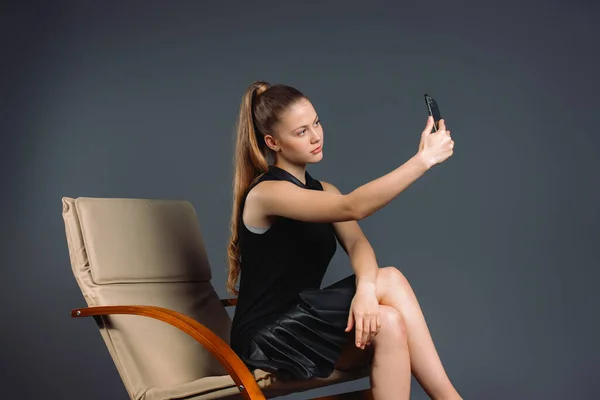 Молодая деловая леди делает селфи на смартфоне, сидя в кресле на сером фоне с боковым пространством. Онлайн работа — стоковое фото