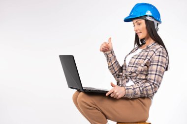 A woman architect in a hardhat, communicates via video on notebook, thumb up gesture, approval or agreement on white. High quality photo