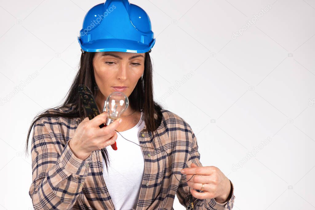 A serious female electrician examines a light bulb in a hard hat on a white background. Breaking gender stereotype. High quality photo