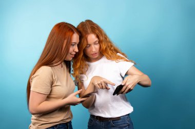 Gündelik giysiler içindeki kızıl saçlı kız arkadaşlar, akıllı telefondaki fotoğraflara göz atmak. Mavi arka planda izole edilmiş. İnsanların yaşam tarzı konsepti. Yüksek kalite fotoğraf