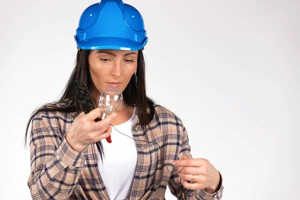 En seriös kvinnlig elektriker undersöker en glödlampa i en hård hatt på en vit bakgrund. Bryta könsstereotypen — Stockfoto
