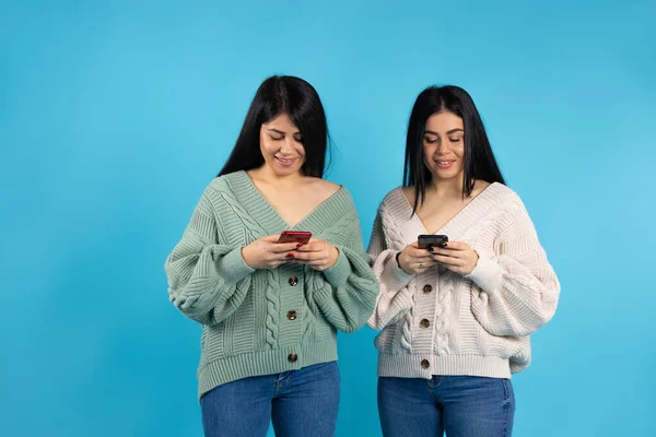 Portrait of cute adorable female twins on a blue background, looking at smartphones in their hands. — Stock Photo, Image