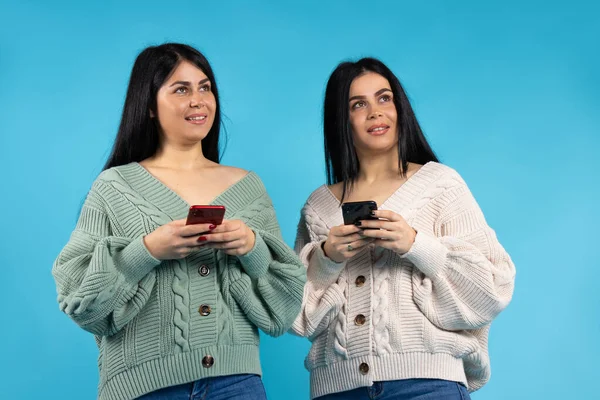 Beautiful twin girls, equally dressed on a blue background, with smartphones in their hands looking up. — Stock Photo, Image