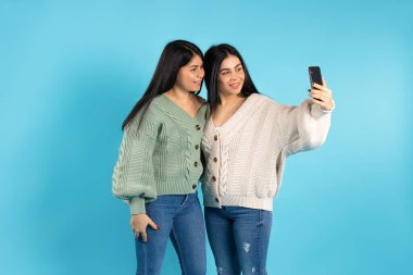İkiz kardeşler gülümseyip cep telefonuyla selfie çekiyorlar. Mavi arkaplan ve boş yan boşluk. Yüksek kalite fotoğraf