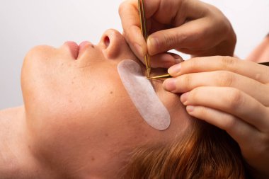 Büyütülmüş fotoğraf. Bir güzellik uzmanının elleri kızıl saçlı genç bir kızın kirpik uzatılması için çalışıyor. Yüksek kalite fotoğraf