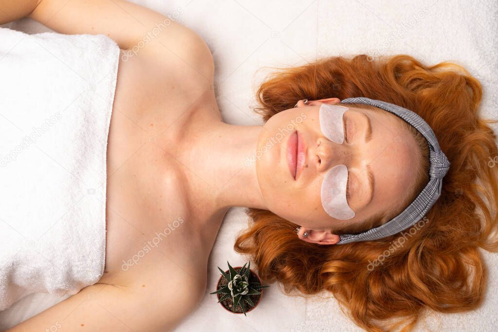 A young red-haired woman wearing a white towel and eye patches, relaxes with closed eyes. Skinimalism and glowing skin. High quality photo