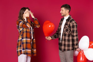 Kırmızı arka planda bir sürpriz bekleyen elleri ile gözlerini kapatan bir kız. Kalp şeklinde balonları olan bir adam. Sevgililer Günü. Yüksek kalite fotoğraf