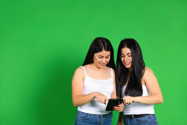 The twin sisters smile and stare at the tablet against a green background with an empty side space. — Stock Photo, Image