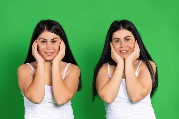 Irmãs gêmeas são bonitos posando com as mãos em seus rostos em um fundo verde, sorrindo e olhando para a câmera. — Fotografia de Stock