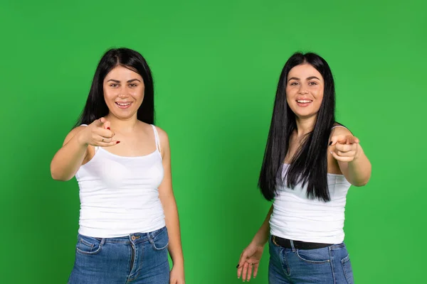 Meninas gêmeas sorridentes apontam para a câmera com um indicador. Fundo verde. — Fotografia de Stock