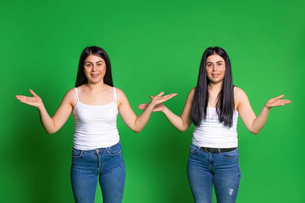 Les jeunes filles jumelles dans les mêmes vêtements redressent leurs mains sur les côtés et regardent la caméra. Fond vert. — Photo