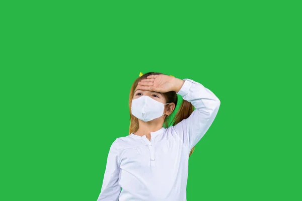 Egy óvodás lány védő maszkban, ráncolja a szemét, fogja a fejét, és zöld háttérrel néz a távolba. Oldaltér. — Stock Fotó