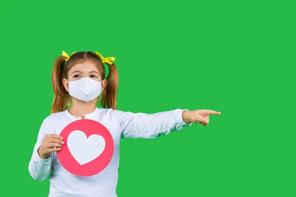 Una niña preescolar con una máscara protectora sostiene un signo con un emoticono rojo del corazón y muestra su dedo índice a un lado. — Foto de Stock