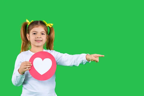 Ett barn på en grön bakgrund med ett hjärta visar pekfingret åt sidan. Sidoutrymme. — Stockfoto