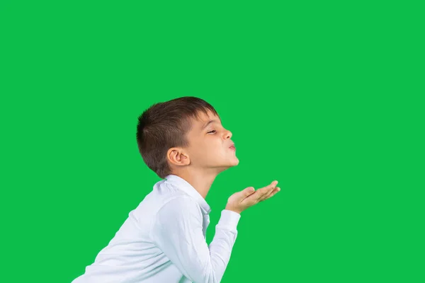 Foto de perfil. El chico sobre un fondo verde envía un beso aéreo. Fondo verde y espacio lateral. Emociones infantiles. — Foto de Stock