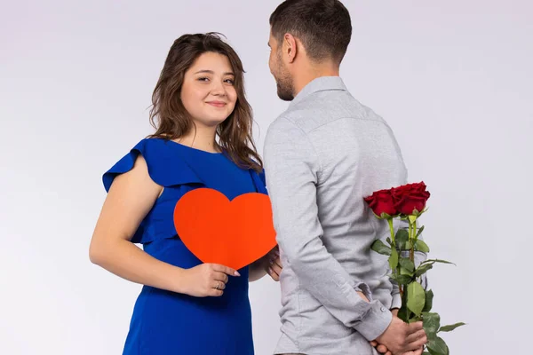 Gri arka planda bir çift. Adam arkasında bir buket kırmızı gül tutuyor. Kartondan kalbi olan kız kameraya bakar.. — Stok fotoğraf