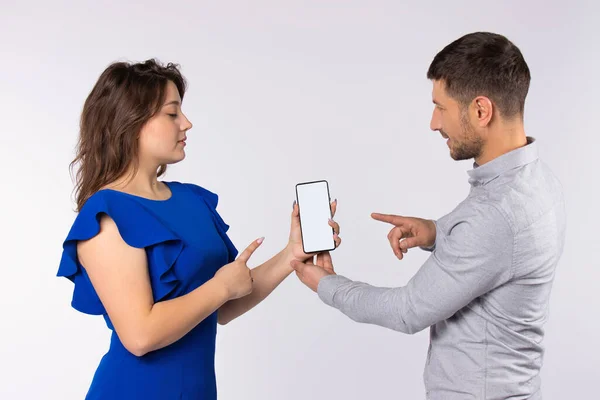Unga par på grå bakgrund håller en smartphone med skärm till kamera, visar det med pekfingret. — Stockfoto