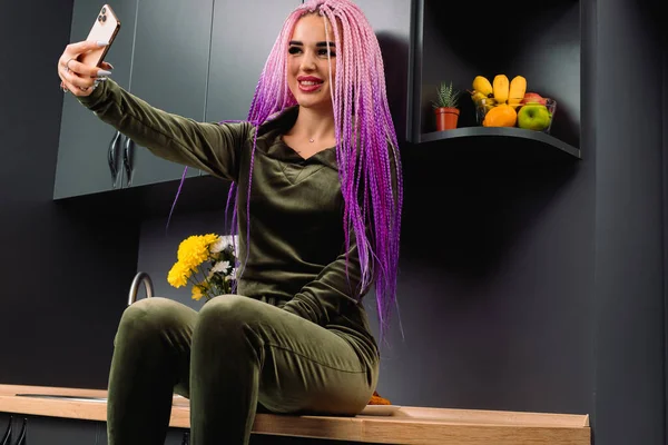 Uma jovem faz uma selfie enquanto se senta em casa na cozinha escalada na mesa. Jovem blogueiro. — Fotografia de Stock