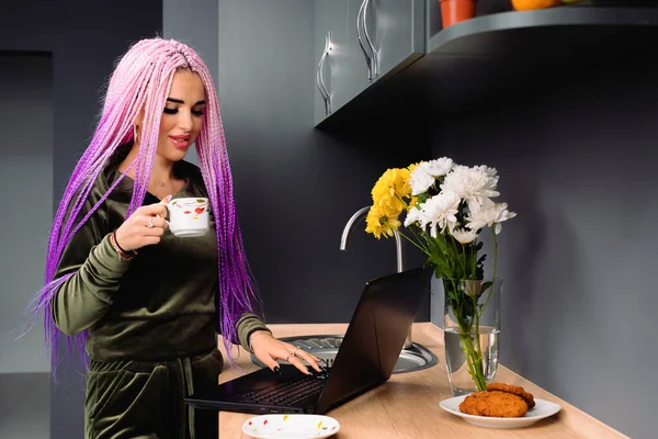 Uma jovem garota moderna em casa na cozinha com uma xícara de café atrás de um laptop está blogando. — Fotografia de Stock