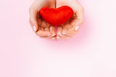 Eller pembe arka planda yumuşak kırmızı pelüş bir kalp tutar. Alttaki boş alan reklam için kullanılır..