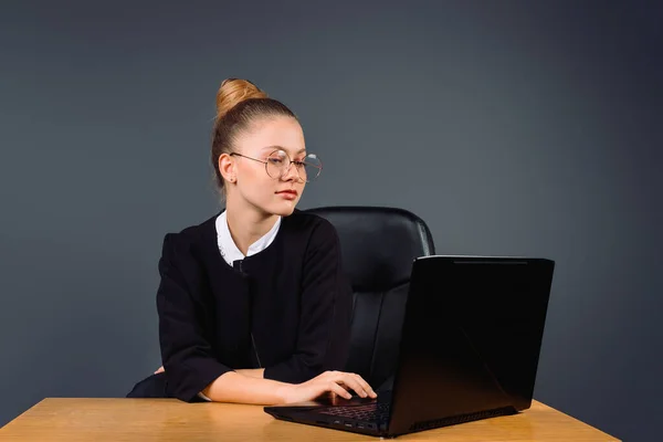 Una donna d'affari carina con gli occhiali si siede in ufficio e tipi su un computer di lavoro. Fondo grigio. — Foto Stock