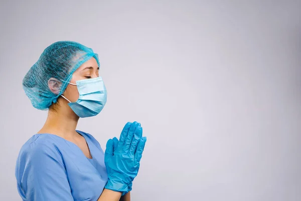 Una doctora con una máscara médica rezando por la salud de pacientes enfermos con los ojos cerrados. Fondo gris. Espacio lateral. — Foto de Stock