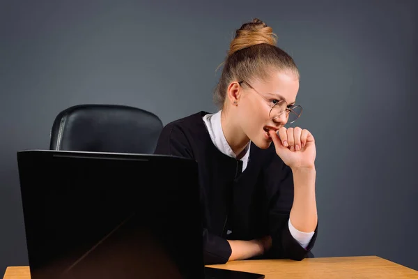 Una donna d'affari con gli occhiali al computer distoglie lo sguardo e si morde le unghie. Stress ed esperienza su sfondo grigio. — Foto Stock