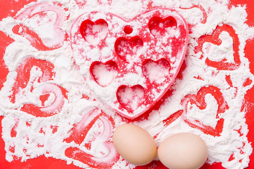 Heart-shaped red silicone mold, eggs and sprinkled flour on a red background.