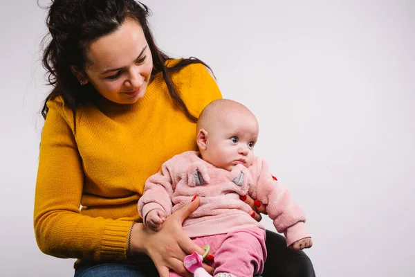 Una niña pequeña en brazos de sus madres mira hacia un lado sobre un fondo gris con espacio lateral vacío. — Foto de Stock