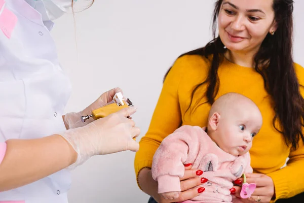 Un médico con una bata médica blanca prepara una pistola perforadora y pendientes médicos. Mamá sonríe, sosteniendo al bebé en sus brazos. Foto recortada — Foto de Stock