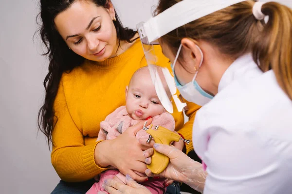 Mamá con la niñera en sus brazos y un médico en una máscara médica y una pantalla facial pandémica con arma perforante contra la propagación del virus. — Foto de Stock