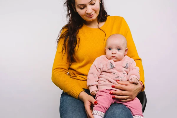 Una niña con grandes ojos azules mira a la cámara mientras está sentada en los brazos de sus madres después de perforar la oreja sobre un fondo gris. — Foto de Stock