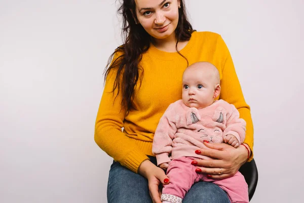 Una joven madre con una sonrisa mira a la cámara sosteniendo a una niñita con piercing en el pendiente sobre un fondo gris con espacio lateral. — Foto de Stock