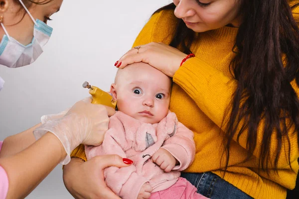 El médico hace agujeros para pendientes para la niña con una pistola perforadora. Mamá sostiene su cabeza. Fondo gris. — Foto de Stock