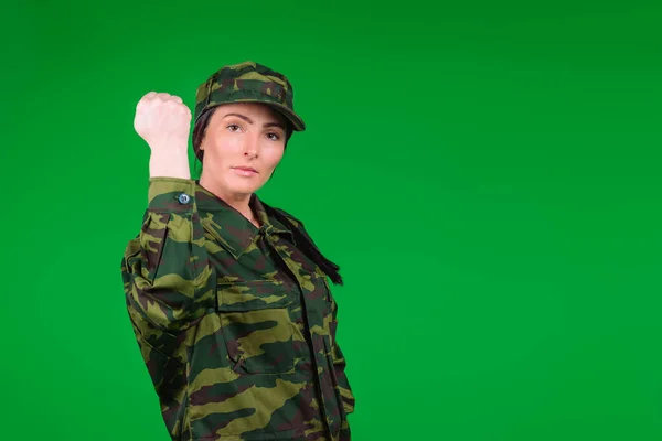 Mujer joven en uniforme militar muestra su puño a la cámara sobre un fondo verde con espacio en blanco. Fuerte valiente soldado femenino — Foto de Stock