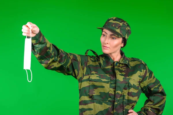 Una mujer seria con uniforme militar sostiene una máscara médica sobre un fondo verde. Neutralidad de género en la pandemia covid 19 — Foto de Stock