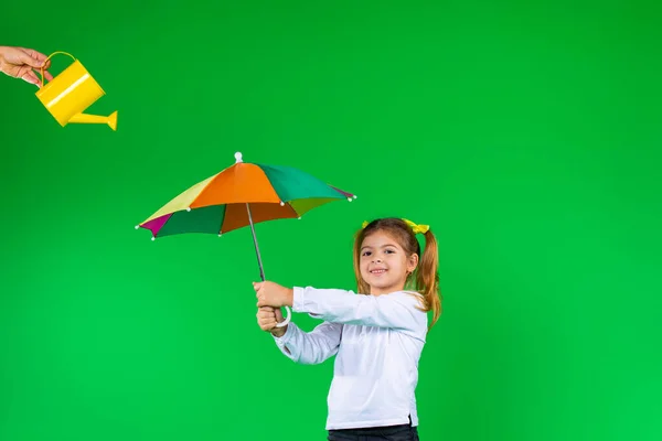 Sonriente niña preescolar posando con un paraguas de color sobre un fondo verde y una regadera amarilla en la parte superior. — Foto de Stock