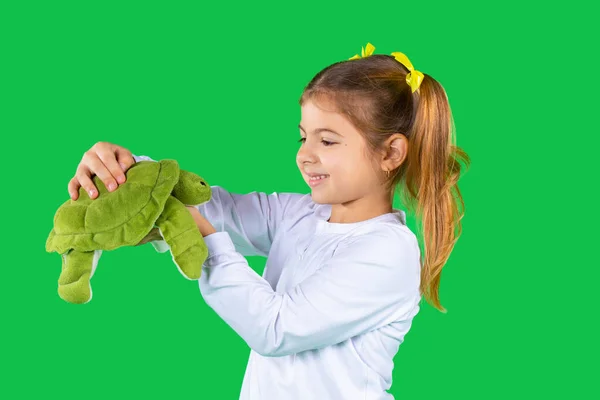 Дошкольная девочка мило улыбается и держит мягкую зеленую черепаху на зеленом фоне. — стоковое фото