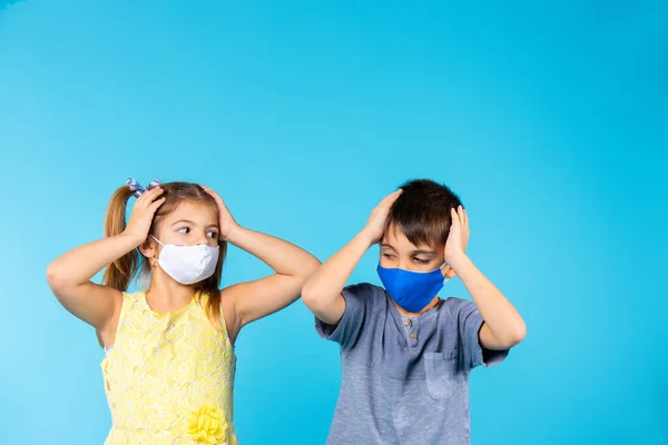 Crianças usando máscaras protetoras seguram suas cabeças com as mãos e olham umas para as outras contra um fundo azul com um espaço superior vazio. Conceito de proteção de vírus. — Fotografia de Stock