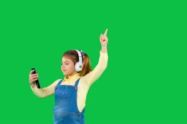스마트폰을 하고 헤드폰을 큰 헤드폰을 끼고 있는 여자 아이가 옆 공간이 있는 녹색 배경에서 검지 손가락을 들고 있습니다.. — 스톡 사진
