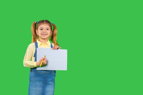Ένα χαριτωμένο μικρό κορίτσι με ένα χαμόγελο κρατώντας ένα λευκό φύλλο σε ένα πράσινο φόντο με κενό χώρο πλευρά για τη διαφήμισή σας — Φωτογραφία Αρχείου