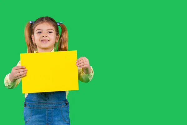 En förskoleblond flicka, leende typ, som håller en lysande banner för reklam. Grön bakgrund och sidoutrymme. — Stockfoto